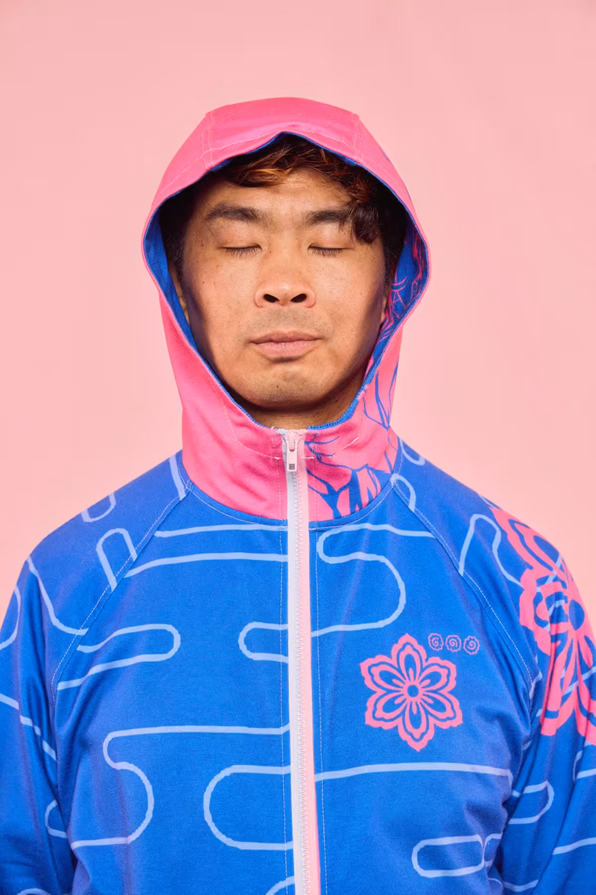 Man with eyes closed wearing a blue and pink hooded jacket with floral patterns against a pink background.