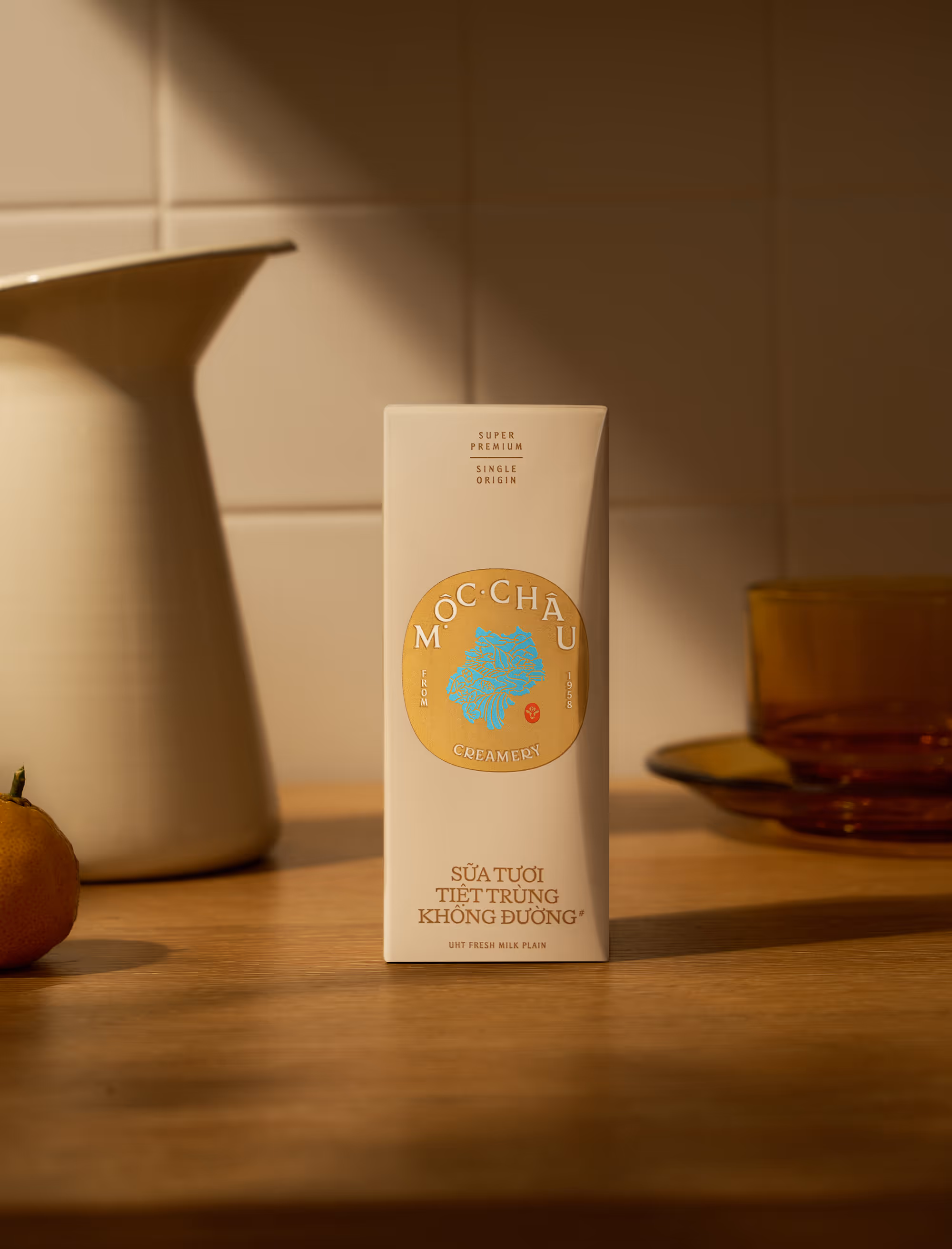 Carton of Moc Chau Creamery UHT fresh plain milk on a wooden table with a white pitcher, an orange, and amber glassware in warm lighting.