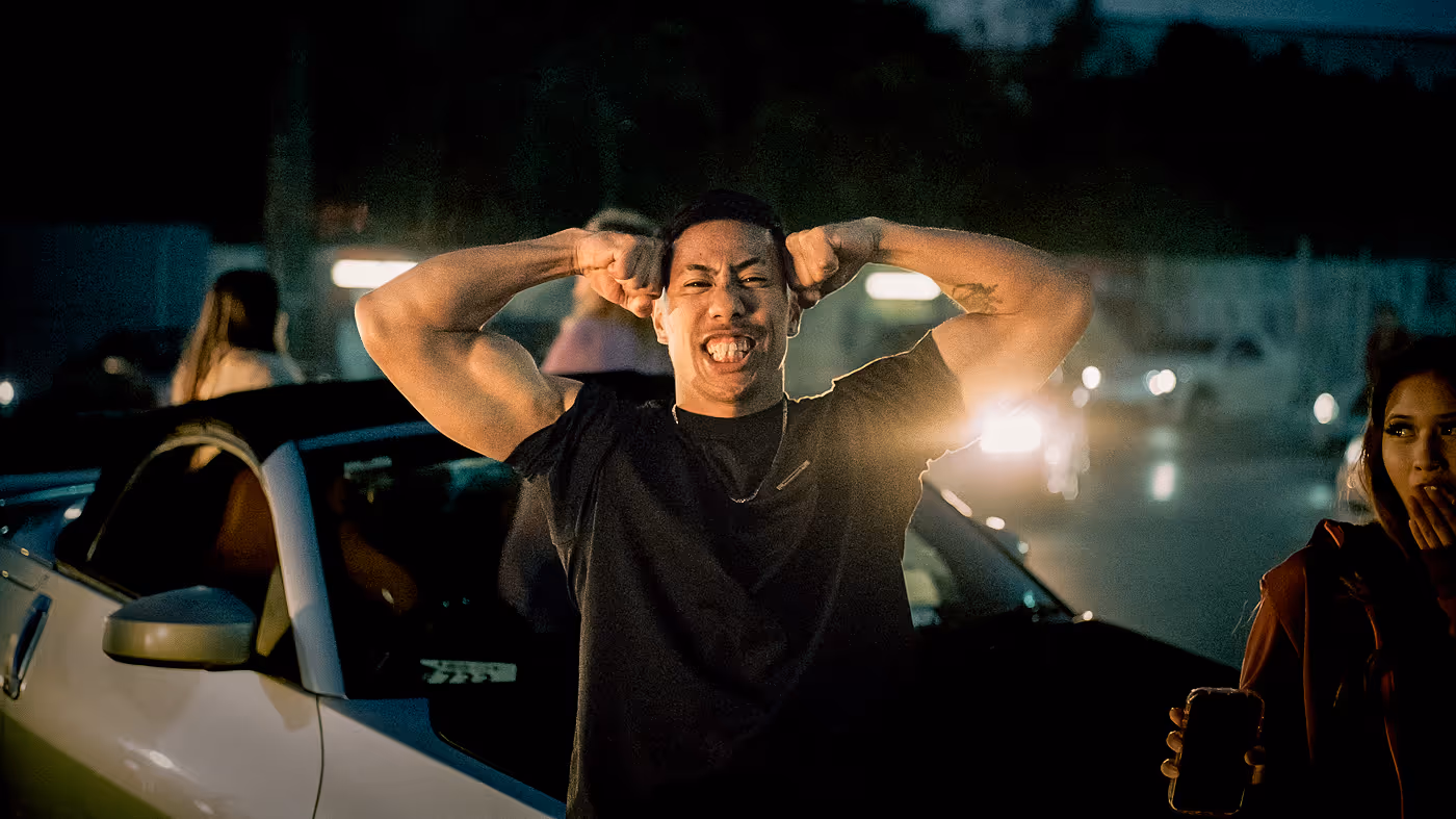 Man flexing his muscles and smiling while standing in front of a white car at night.